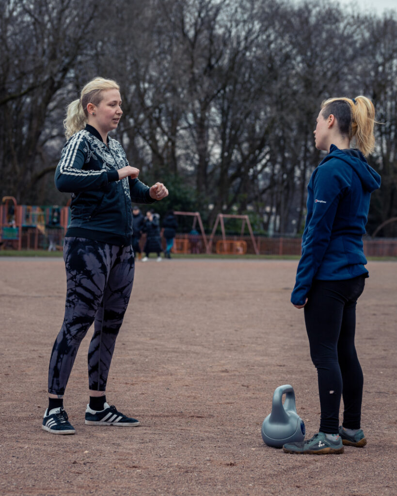 A wide shot of Dylan, a blonde non-binary person, and a woman
with bright orange hair. They are facing each other and there is a
kettlebell at the woman's feet. Dylan has raised arms and is explaining an
exercise as the woman listens. They are in an outdoor park environment with
a sandy floor and a playground in the background and trees behind that.