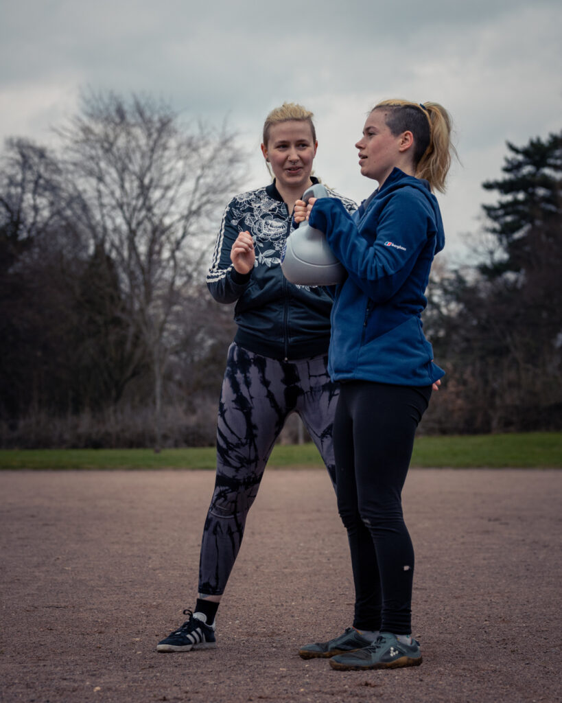 A wide shot of Dylan, a blonde non-binary person, and a woman
with bright orange hair.  The woman is facing side on to the camera and
holding a kettlebell up close to her chest as Dylan stands to her side,
face on towards the camera smiling and looking at the woman as she is about to begin her exercise.