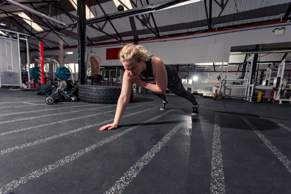 Wide angle shot of Dylan, a blonde non-binary person, in the middle of a
gym environment. They are mid way through a shoulder tap in a straight
armed plank position with a look of focus on their face. In the background
there are squat racks, barbells and a tyre.